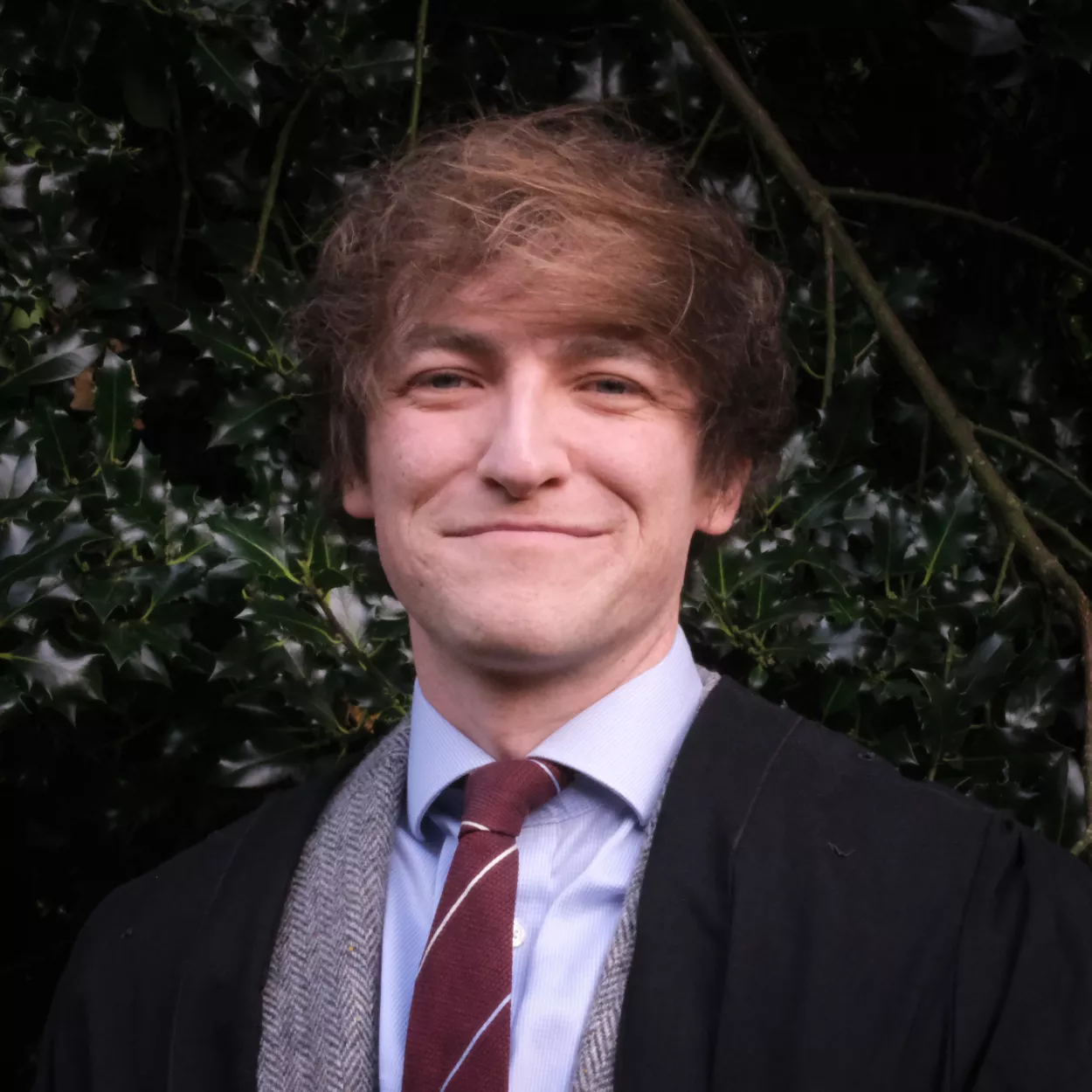 Miles is smiling at the camera. He is wearing a grey suit, maroon tie and black gown.